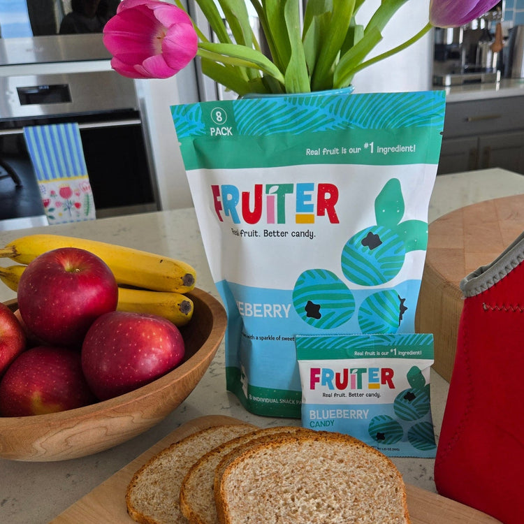 Fruiter all-natural dried blueberry candy snack bag on a kitchen counter next to a child's lunch box with other healthy lunch options. Fruiter candy comes in sweet, sweet and sour, and super sour varieties.